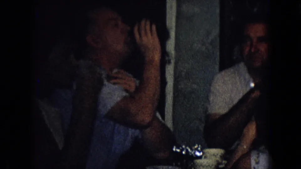 A man drinks from a glass at a dark indoor party in 1965 New Jersey, with seated adults and tableware partly visible around him.