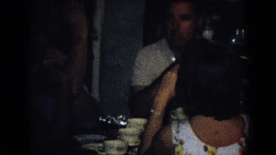 Adults at a 1965 New Jersey home gathering sit near cups or bowls on a table, with one figure partially obscured by the handheld camera framing.
