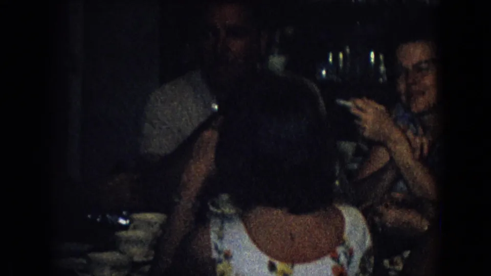 A woman in patterned clothing gestures or points while seated near other adults at a dim 1965 New Jersey indoor party table.