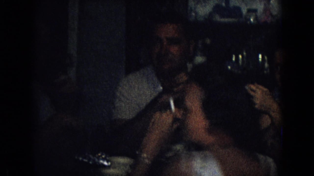 In this 1965 New Jersey home-movie frame, adults sit close together in a dim private interior while a man in a light shirt holds a glass near the camera.