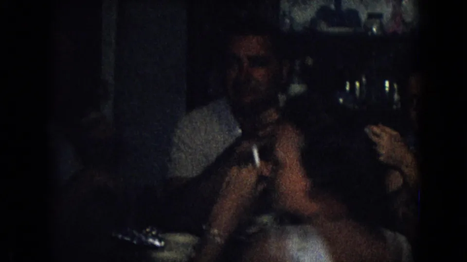 In this 1965 New Jersey home-movie frame, adults sit close together in a dim private interior while a man in a light shirt holds a glass near the camera.