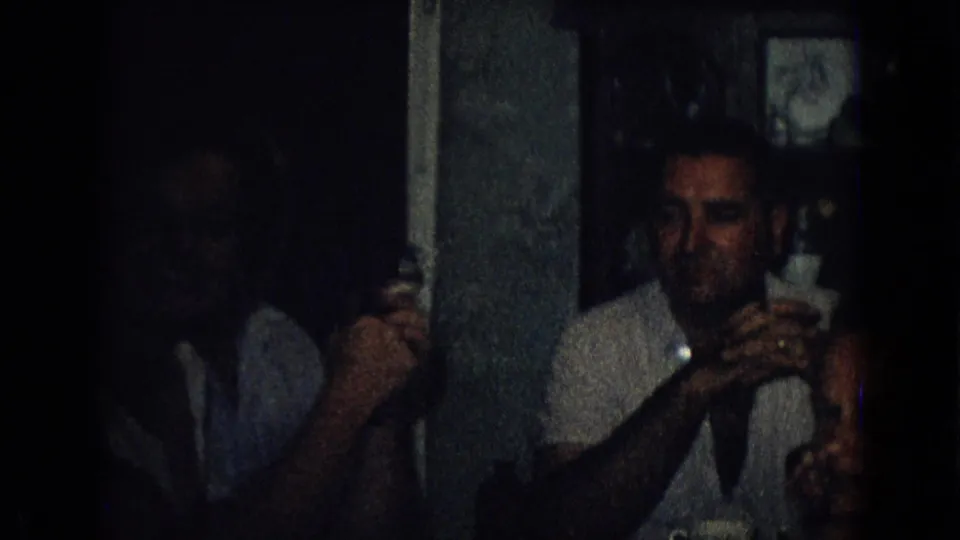 A seated adult man in a light shirt appears beside a vertical interior partition in a dark 1965 New Jersey private gathering, with a drink in his hands.