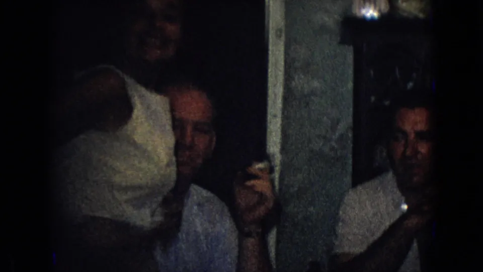 Two adult men are visible in a shadowed private interior from the 1965 New Jersey home movie, with one man partly obscured and another holding a small glass.