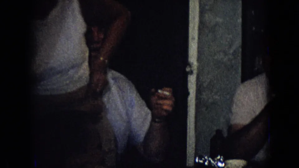 Adults sit close together in a dark private interior in this 1965 New Jersey frame, with a foreground figure partially obscuring seated people and a man at the right edge.