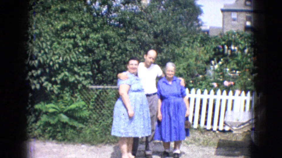 Three adults pose together in a bright New Jersey backyard in 1965, with two women in blue dresses and a man in a white T-shirt standing before greenery and a white picket fence.