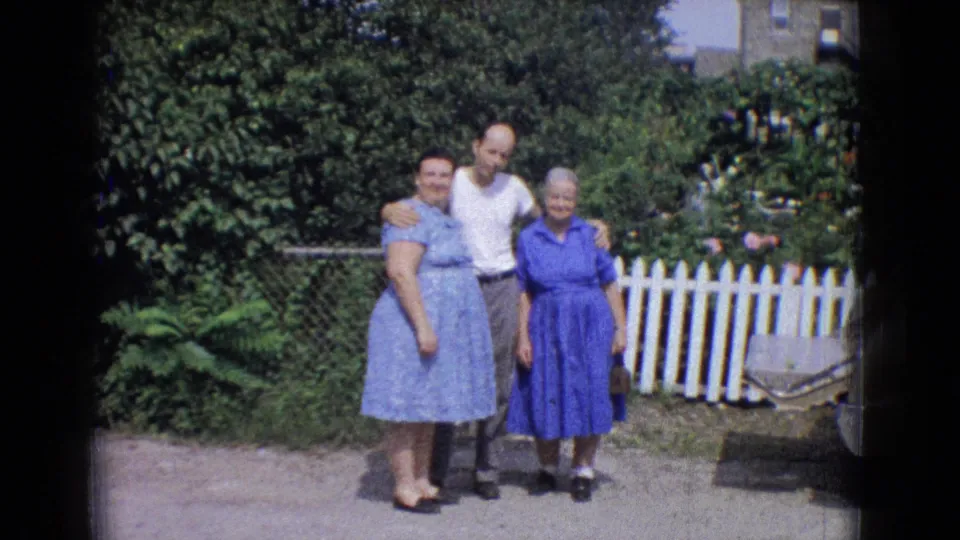 Two women in blue dresses and a man in a white T-shirt stand closely together for the camera in a 1965 New Jersey backyard with shrubs and a white fence behind them.