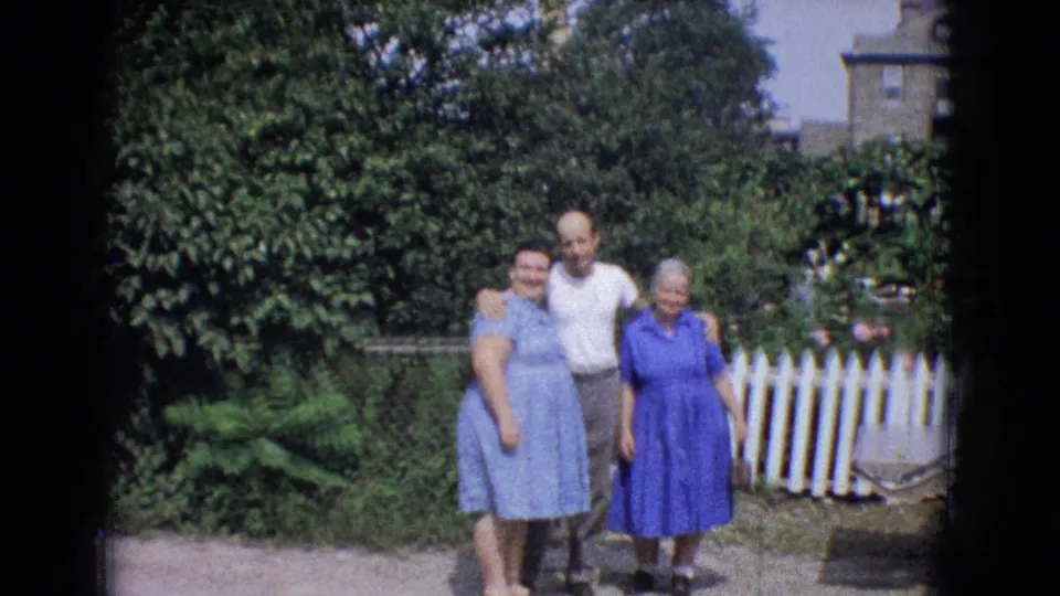 The three adults remain posed in the sunny backyard, framed by leafy bushes, a white picket fence, and partial residential structures in this 1965 New Jersey 8mm frame.
