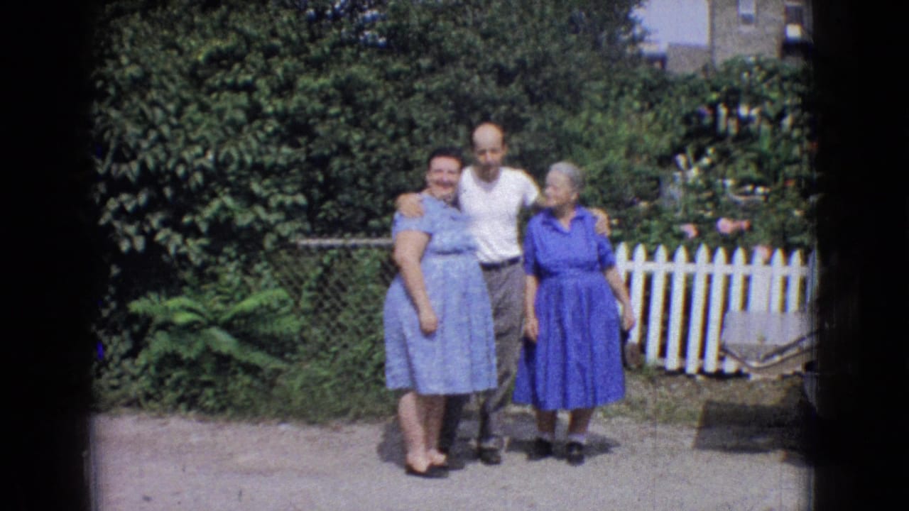 In 1965 New Jersey home-movie footage, a man in a white T-shirt stands between two women in blue dresses as they pose in a backyard near fences and foliage.