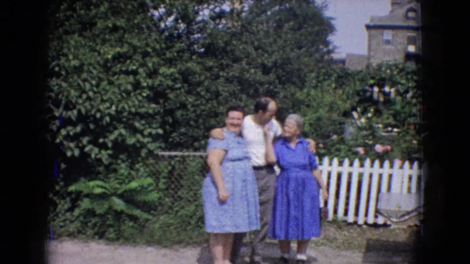 The backyard pose continues as the man stands with his arms around two women in blue dresses, with a white picket fence and dense greenery behind them.