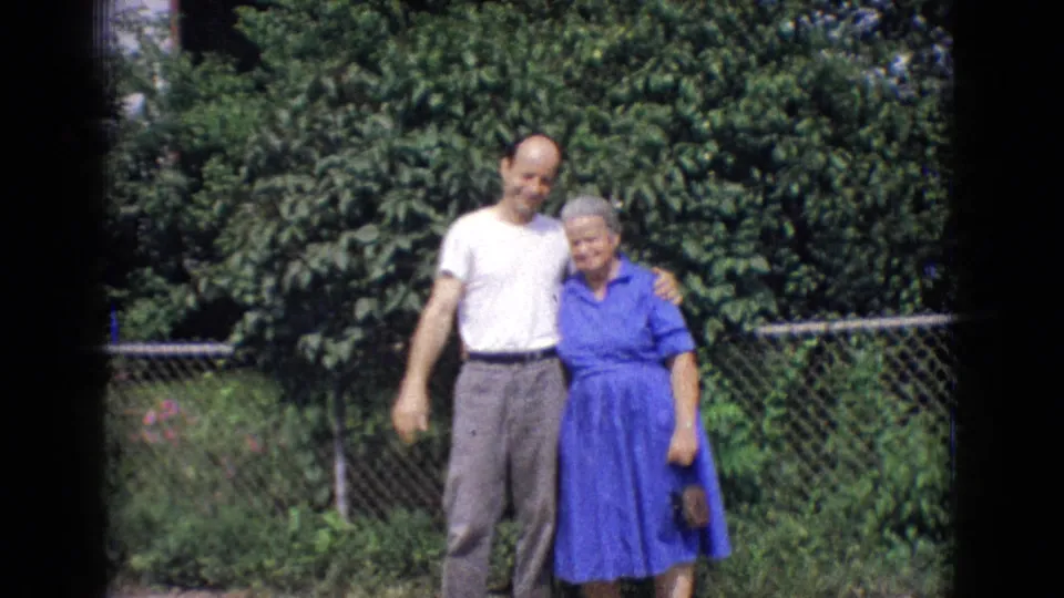 In the later part of the 1965 backyard scene, the man and an older woman in a blue dress pose together against a chain-link fence and garden foliage.