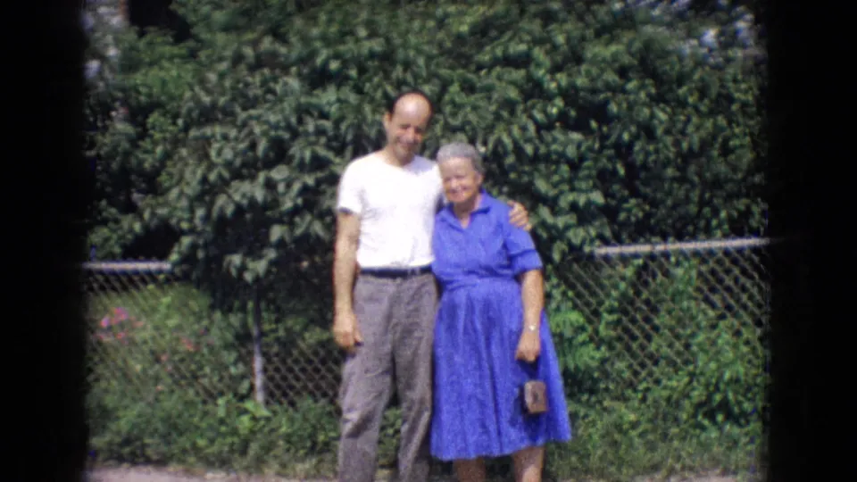 The man and older woman continue posing in the New Jersey backyard, standing close in front of foliage and a chain-link fence with soft 8mm color texture.