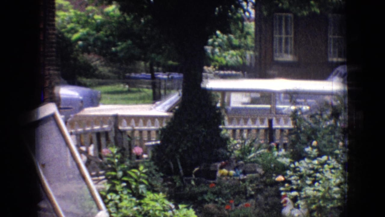 1965 New Jersey 8mm frame looking past a leafy garden and white picket fence toward a residential street with a brick house and classic cars partly visible.