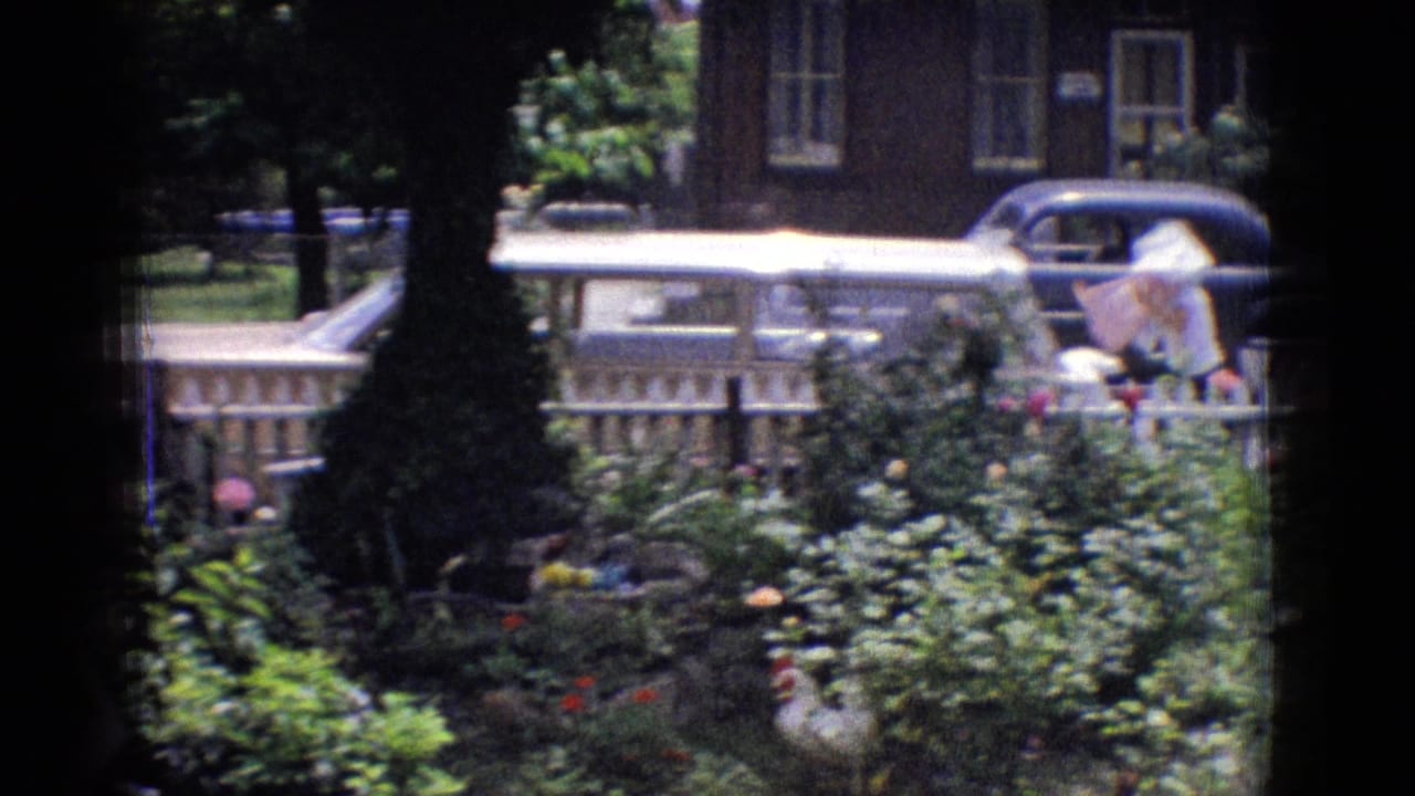 1965 New Jersey home-movie frame with a garden foreground, white fence, brick residential building, and a parked classic car partly visible across the street.