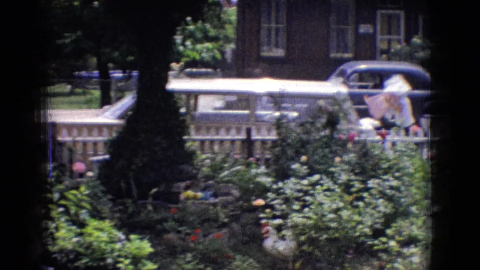 1965 New Jersey home-movie frame with a garden foreground, white fence, brick residential building, and a parked classic car partly visible across the street.