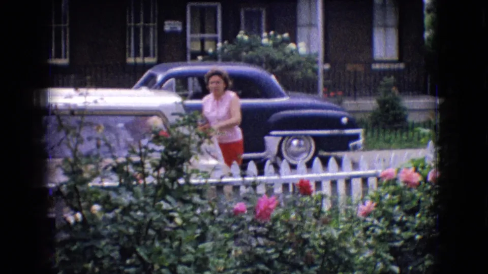 1965 New Jersey 8mm frame of a woman standing near a blue classic sedan behind a white picket fence, with roses, greenery, and a brick residential facade behind her.