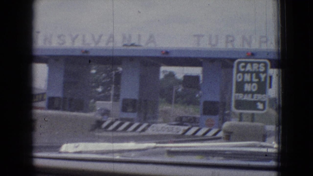 1965 8mm home-movie frame from an in-car approach toward a blue toll plaza, with a cars-only no-trailers sign and roadway barriers visible ahead.