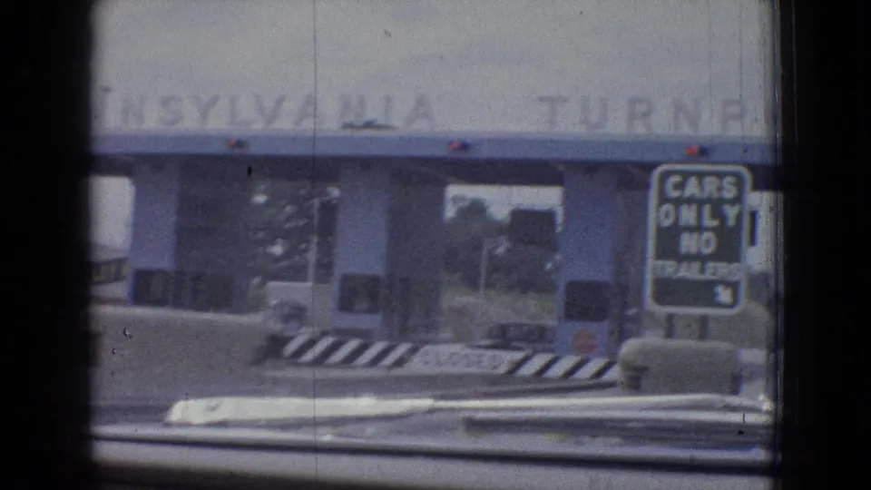 1965 8mm home-movie frame from an in-car approach toward a blue toll plaza, with a cars-only no-trailers sign and roadway barriers visible ahead.