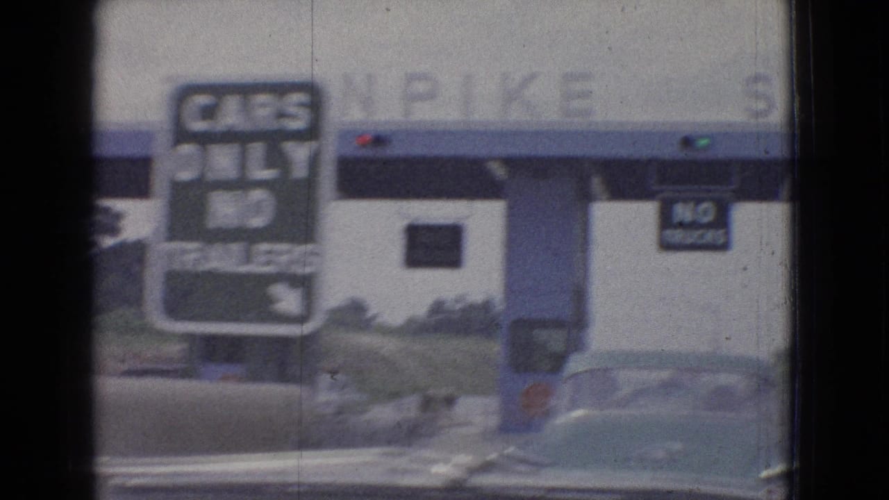 In-car 1965 road-travel frame showing a toll booth lane, cars-only signage, and a nearby vehicle as the camera faces the plaza through the windshield.