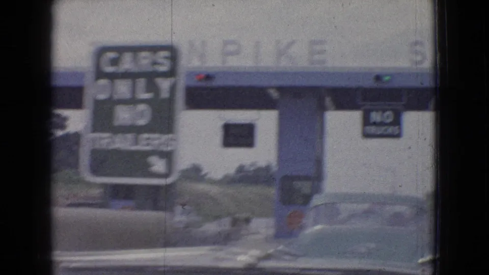 In-car 1965 road-travel frame showing a toll booth lane, cars-only signage, and a nearby vehicle as the camera faces the plaza through the windshield.