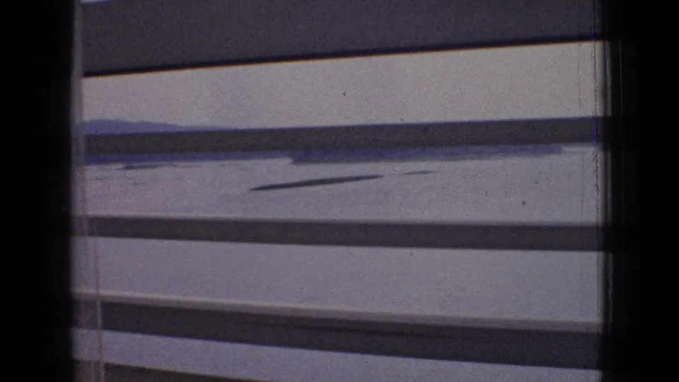 Soft 8mm frame from inside the vehicle, with dark window framing, roadway surface, lane edge, and low open landscape visible beyond the toll area.