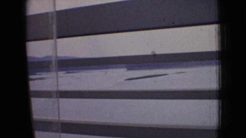 Final 1965 in-car frame looking past window framing and pavement toward a flat, open roadside landscape, with the toll plaza no longer prominent.