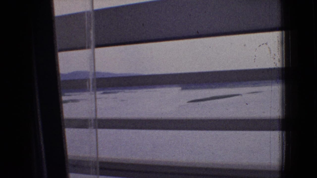 1965 New Jersey 8mm train-window view through horizontal blinds, with pale sky, calm water, and distant low hills visible beyond the dark window frame.