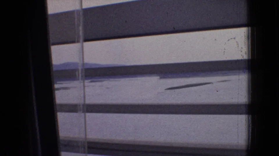 1965 New Jersey 8mm train-window view through horizontal blinds, with pale sky, calm water, and distant low hills visible beyond the dark window frame.