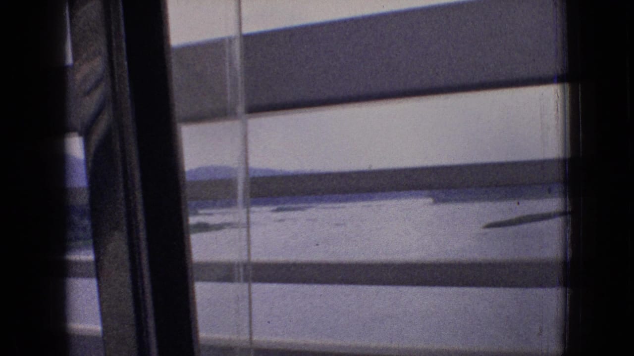 Blurred landscape seen through train-window blinds with water and low hills beyond.