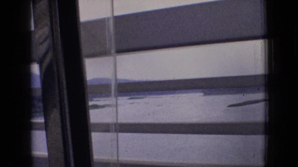 1965 New Jersey train-window view in 8mm, showing horizontal blinds, a pale lake surface, distant hills, and dark interior framing as the train moves.
