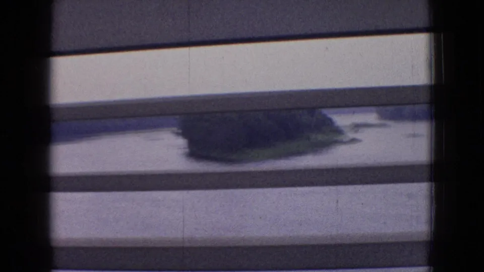 1965 New Jersey home-movie frame from a moving train, with horizontal blinds crossing the view of water, shoreline, and a dark low landform.