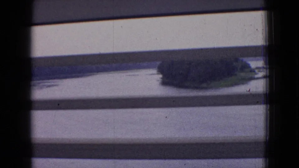 1965 New Jersey 8mm travel view through train-window blinds, with calm water, a small wooded shoreline or island, and soft distant landscape detail.