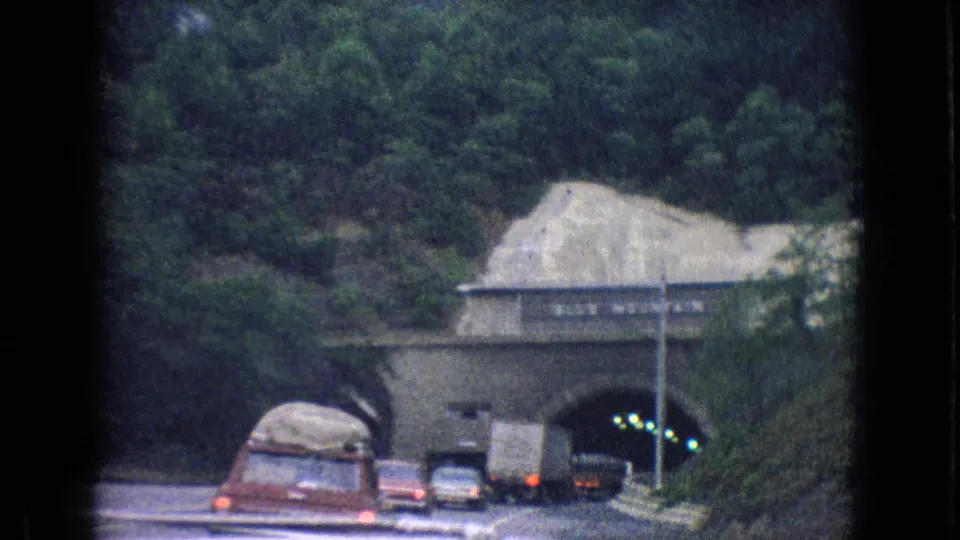Traffic, including a red car, enters a tunnel under a wooded rocky approach in the 1965 New Jersey 8mm road-trip footage.