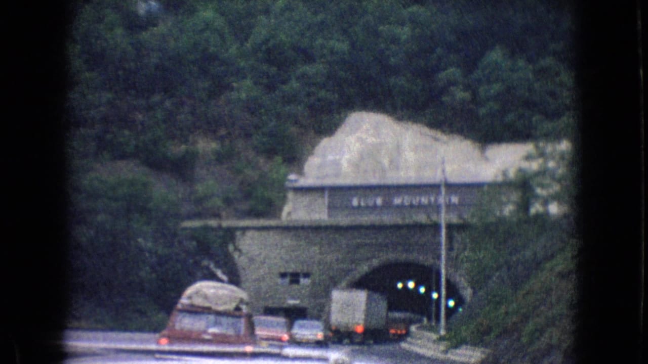 1965 New Jersey 8mm frame showing traffic approaching a tunnel entrance marked Blue Mountain, with forested slopes and a road sign partially visible around the portal.