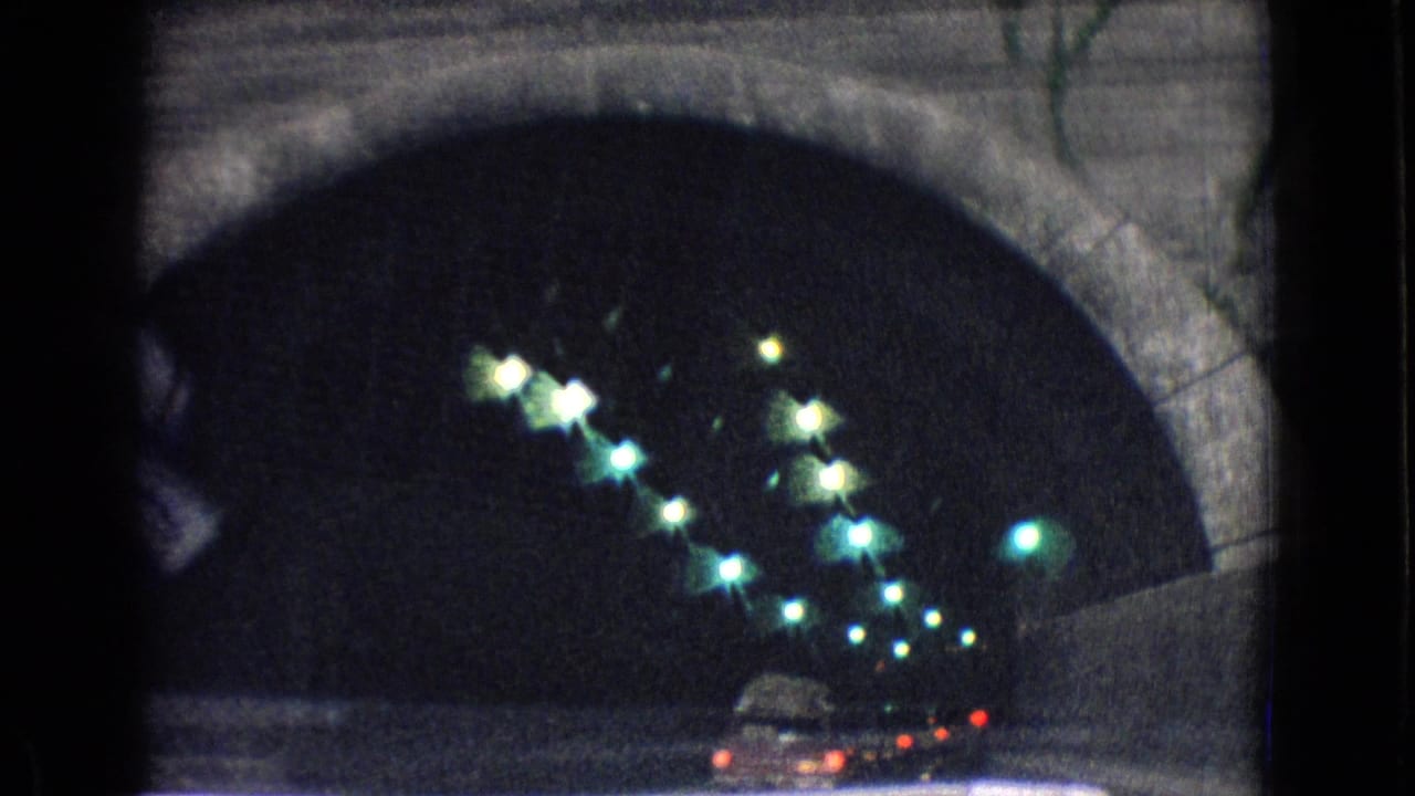 Interior view of the New Jersey mountain tunnel from a moving vehicle, with arched concrete walls, greenish overhead lights, and car tail lights ahead.