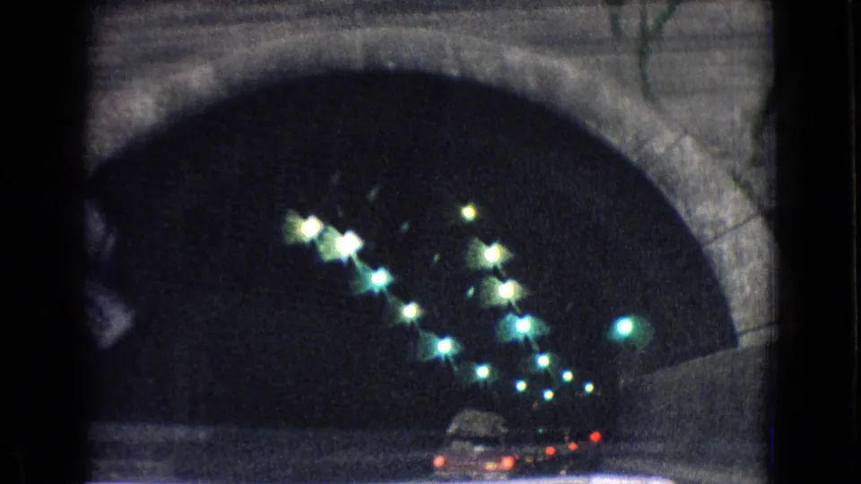 Interior view of the New Jersey mountain tunnel from a moving vehicle, with arched concrete walls, greenish overhead lights, and car tail lights ahead.