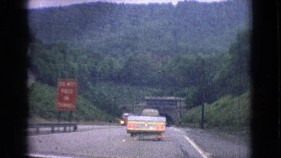 Passenger-view frame from the 1965 New Jersey road trip showing a light-colored car on the roadway near the tunnel area, with signage, guardrails, trees, and hazy hills in the distance.