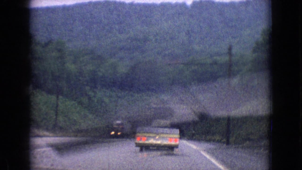 A pale station wagon travels ahead on a winding wooded road in 1965 archival home-movie footage identified as New Jersey.