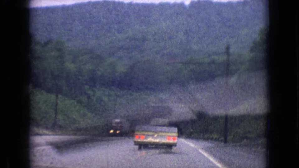A pale station wagon travels ahead on a winding wooded road in 1965 archival home-movie footage identified as New Jersey.