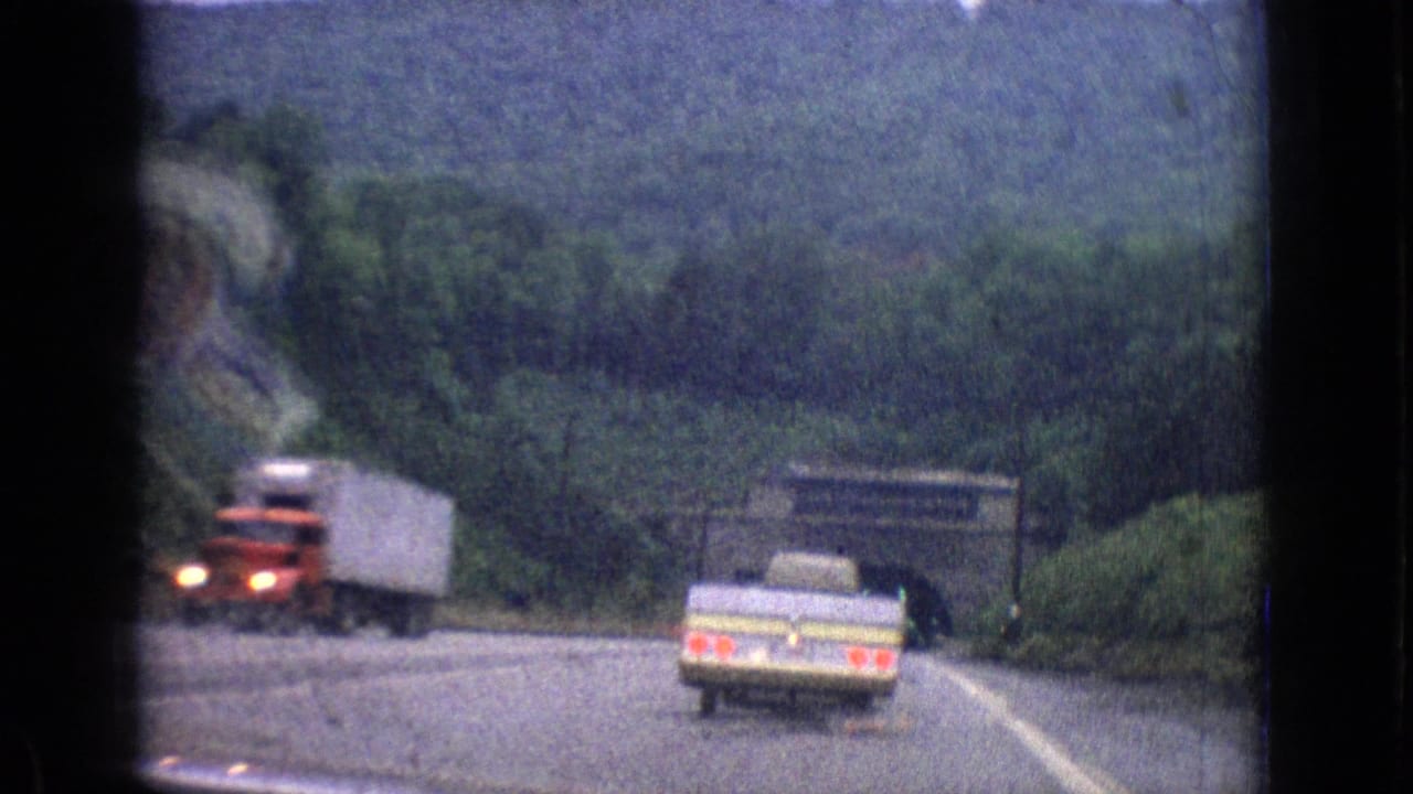 A red semi-truck passes near a pale station wagon on a wooded highway approach in the 1965 road-trip clip.