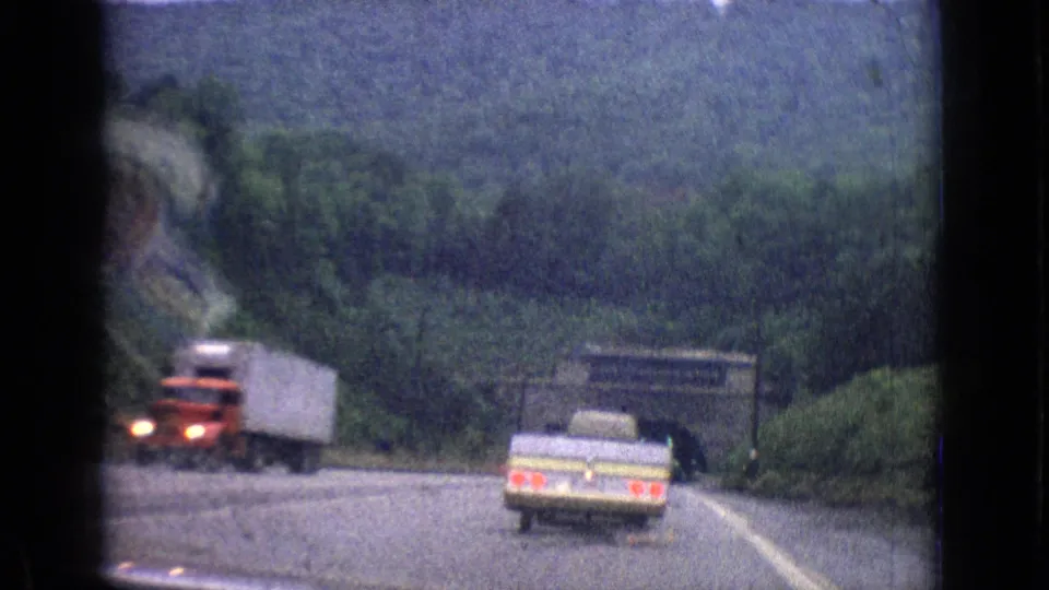 A red semi-truck passes near a pale station wagon on a wooded highway approach in the 1965 road-trip clip.