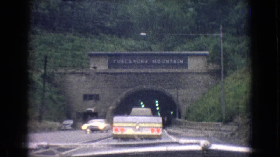 The camera follows a pale station wagon toward a stone-faced tunnel portal on a wooded highway, with overhead lettering visible but not fully relied on for identification.