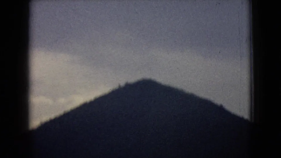 1965 New Jersey home-movie frame with a large dark hill silhouette in the foreground and a muted cloudy sky above.