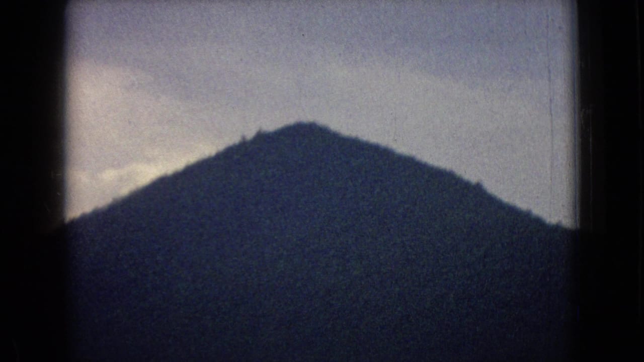 1965 New Jersey 8mm home-movie frame showing a dark mountain silhouette beneath a pale sky, viewed through the edge of a moving vehicle window.