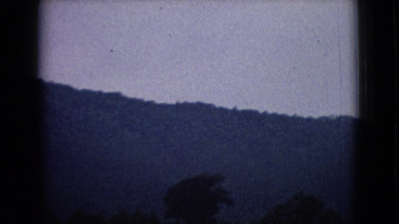 1965 New Jersey road-view frame with a distant mountain ridge, pale sky, window framing, and a blurred dark foreground shape from the moving vehicle.