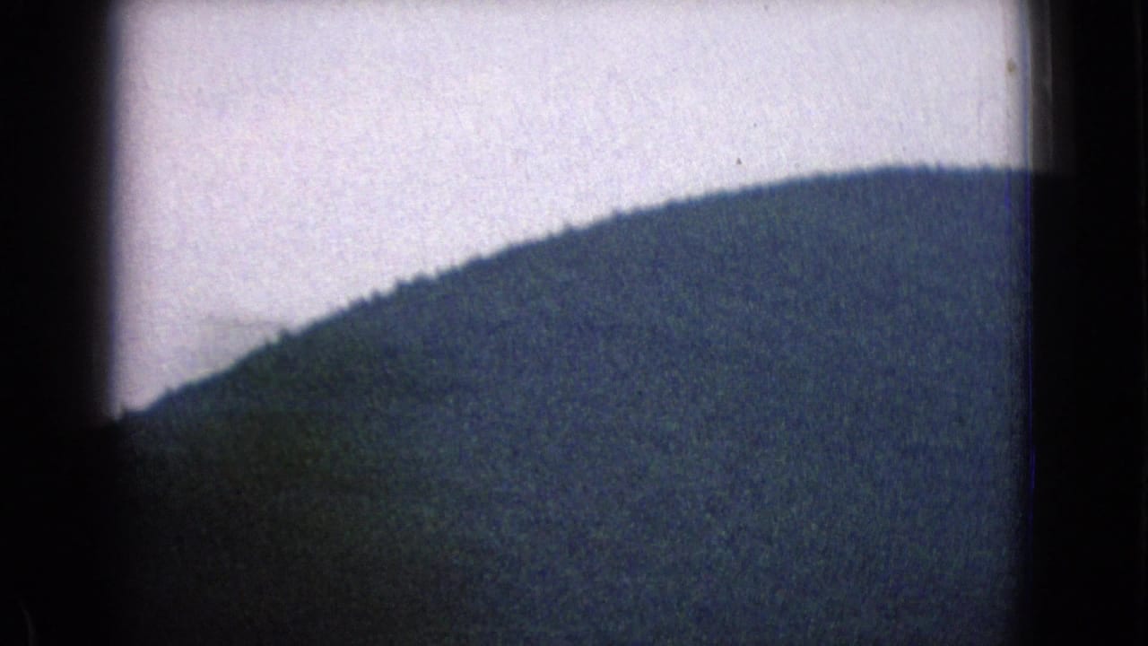 Soft 8mm view of a rounded Sideling Hill landscape beneath a pale sky, partly obscured by dark frame edges from the moving-car footage.