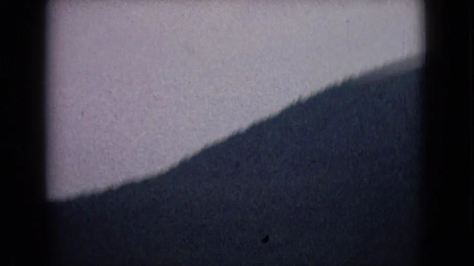 Dim 1965 8mm frame showing the Sideling Hill slope and pale sky, with the landscape partially lost in shadow and film grain.