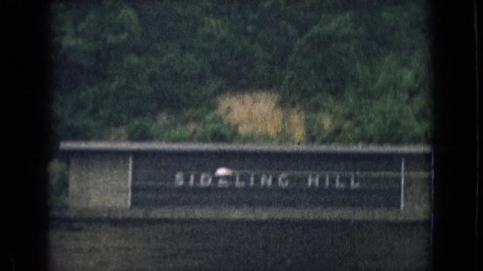 1965 8mm moving-car view of a Sideling Hill sign mounted on a concrete roadside barrier, with green trees and a hillside rising behind it.