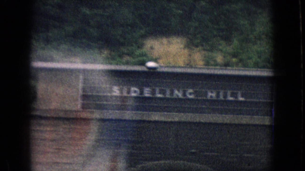 Sideling Hill sign on a roadside structure below a wooded hillside.