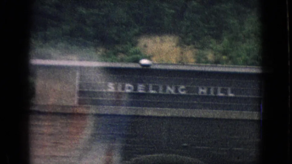 1965 8mm frame showing a Sideling Hill sign on a dark roadside bridge or tunnel structure with trees and hillside above, based on the supplied clip metadata.