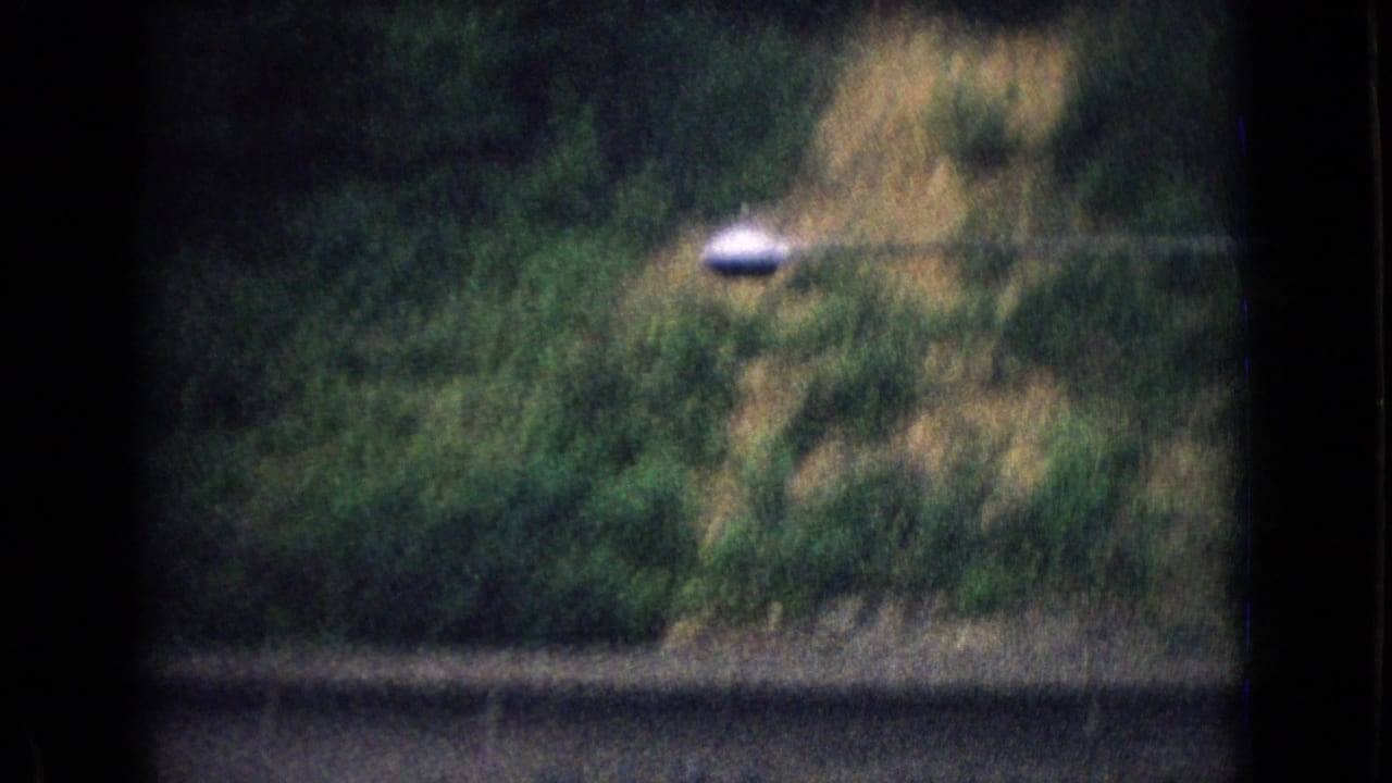 1965 New Jersey 8mm frame showing blurred roadside greenery, a dark foreground barrier, and a wooded hillside seen from a moving vehicle.
